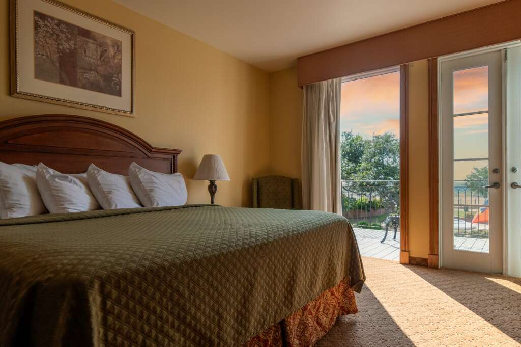 Hotel room with a king-sized bed, open door to a patio, and a view of trees and a cloudy sky.