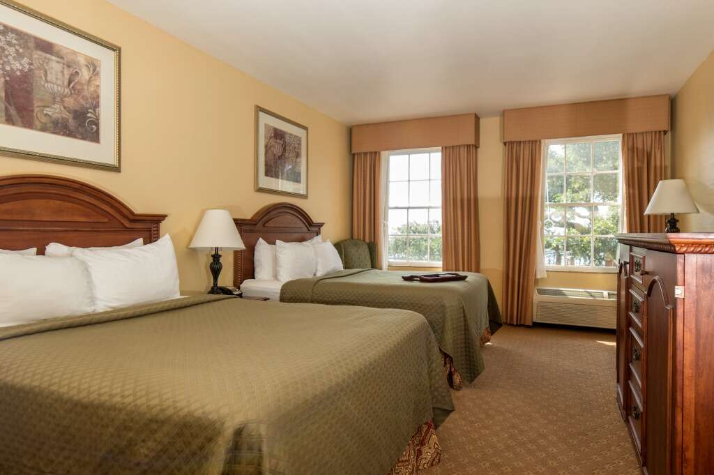 A hotel room with two queen beds, tan walls, and two windows with curtains.