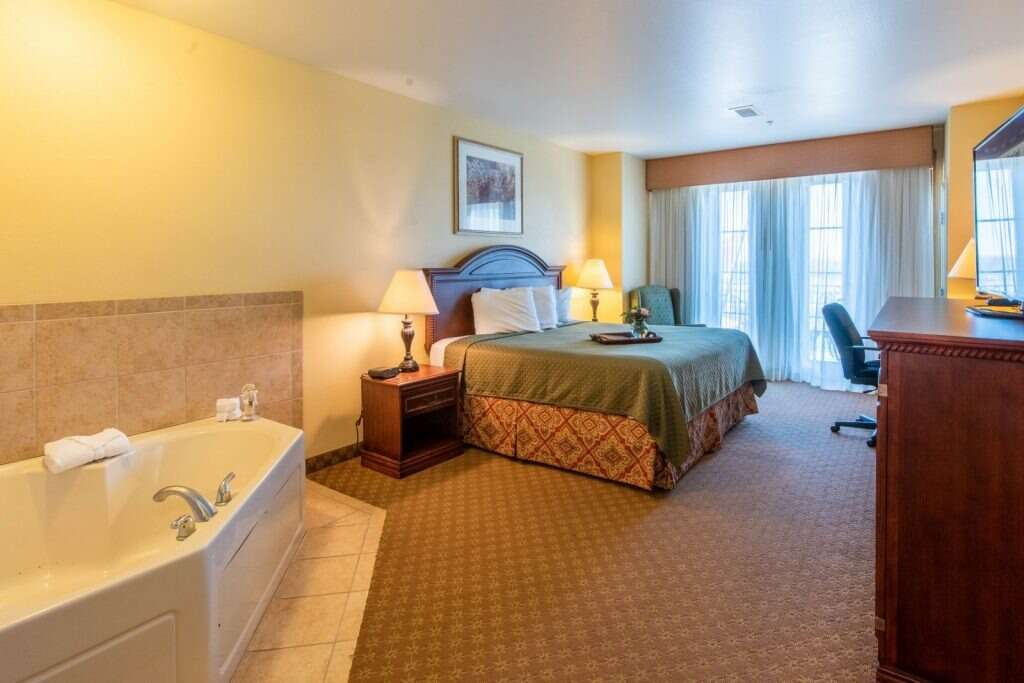 Hotel room with jacuzzi, bed, desk. Yellow walls, beige carpet, and sliding door to balcony.