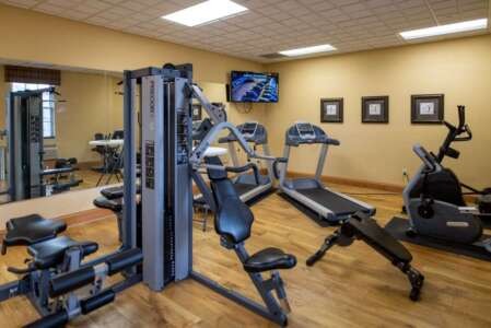Gym with weight machine, treadmill, stationary bike, and TV. Beige walls, wooden floor, and large mirror.