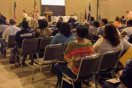 Audience at a conference listens to a speaker at a podium in a large hall. Flags are visible.
