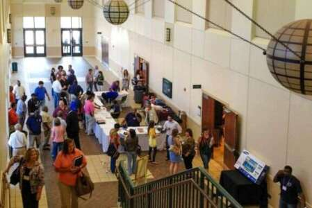 People gathering in a bright, spacious lobby; some at tables. A staircase is visible.