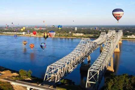 Hot air balloons float over a bridge spanning a river, with a town in the background.