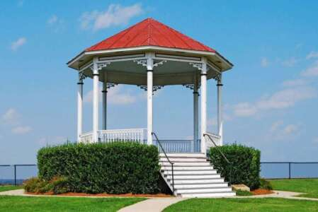 White gazebo with red roof atop steps, framed by hedges, on green grass under blue sky.