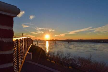 Sunset over a river, framed by a brick wall and bridge. Orange sun reflects on water.
