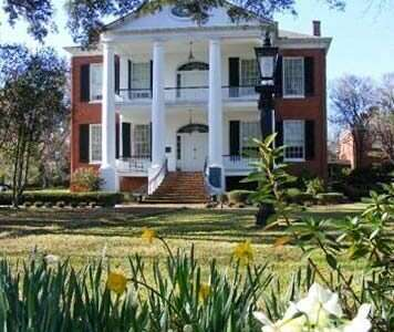 Two-story brick mansion with white columns, black shutters, and a green lawn with daffodils.