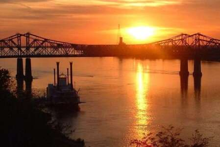Sunset over a river with a bridge and a boat; warm orange and gold colors.