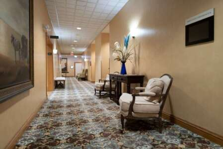 Hotel hallway with floral carpet, beige walls, chairs, and artwork.